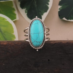 May include: A silver ring featuring a large, oval turquoise stone. The stone is set in a rope-style bezel and has visible veining. The ring has a triple-band design, adding to its unique style. The ring is displayed on a dark wooden surface.