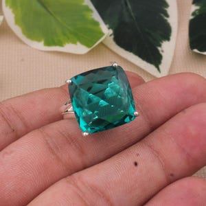 May include: A square-cut, teal-colored gemstone ring. The large, faceted stone is set in a silver band. The ring is held in a hand, with a neutral background of leaves and fabric.