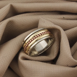 May include: A silver ring with gold and copper bands. The ring has a wide band with a rotating centre section. The ring is set against a neutral-toned fabric background, highlighting its metallic details.
