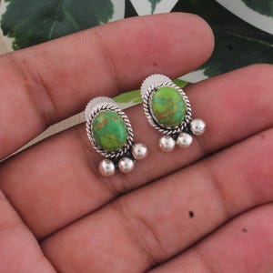 May include: A pair of stud earrings featuring oval-shaped green stones with brown matrix patterns. Each stone is set in a twisted silver frame with three small silver spheres attached at the base, creating a detailed and textured appearance.