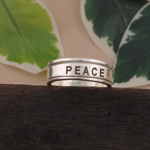 May include: A silver ring featuring the word "PEACE" engraved around the band. The ring is displayed against a backdrop of green and white leaves and a dark wooden surface. The ring's design is simple and elegant.