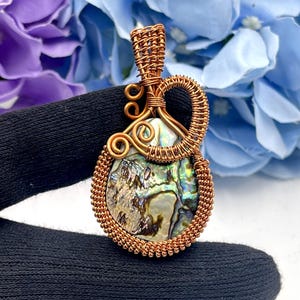 Könnte beinhalten: Abalone-Muschel-Anhänger, umwickelt mit Kupferdraht. Die schillernde Schale ist in einem detaillierten, handgefertigten Drahtdesign eingeschlossen. Der Anhänger wird vor einem Hintergrund aus lila und blauen Blumen gehalten.