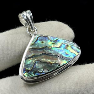 Könnte beinhalten: Abalone-Muschel-Anhänger mit silberfarbenem Metallrahmen. Die schillernde Muschel zeigt Farbtöne von Blau, Grün und Gold. Der Anhänger hat eine dreieckige Form und eine Schlaufe für eine Kette.