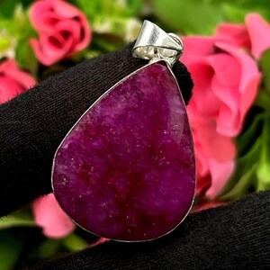 Könnte beinhalten: Ein tropfenförmiger Anhänger mit einem leuchtenden, tiefmagentafarbenen Edelstein, der in einem silberfarbenen Metall gefasst ist. Der Anhänger wird vor einem Hintergrund aus rosa Rosen und grünem Laub präsentiert.