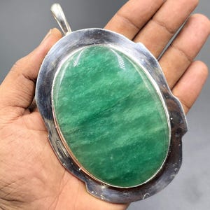 Puede incluir: Colgante grande, de forma ovalada, con una piedra verde pulida y borde de metal plateado. La piedra tiene un aspecto veteado natural. El colgante tiene un lazo en la parte superior para una cadena.