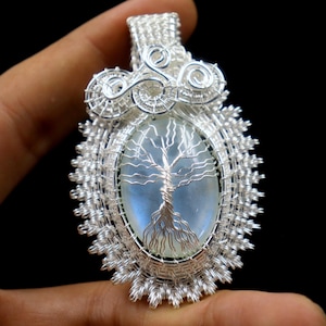May include: A silver-colored pendant featuring a wire-wrapped design. The pendant has an oval-shaped, light blue stone with a wire tree design in the center. The pendant is held by a hand against a black background.