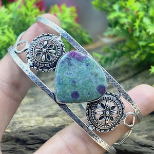 Könnte beinhalten: Ein silberfarbenes Armreif-Armband mit einem großen, herzförmigen grünen Stein mit roten Einschlüssen. Das Armband hat zwei dekorative kreisförmige Elemente mit floralen Mustern.