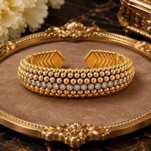 May include: A gold cuff bracelet featuring a textured surface of small, uniform spheres arranged in multiple rows. A single horizontal line of clear, round-cut stones is embedded in the centre of the band, which has an open back design.