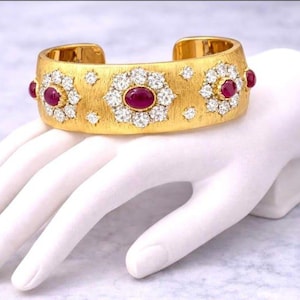 Puede incluir: Un brazalete ancho de oro con textura cepillada, que presenta tres prominentes rubíes rojos de corte ovalado rodeados por grupos de diamantes blancos redondos, con pequeños detalles de diamantes adicionales dispersos por la superficie.