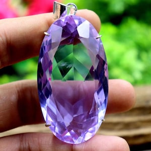 Puede incluir: Un colgante de amatista ovalado y facetado. La gema púrpura tiene una fianza plateada. El colgante se sostiene sobre un fondo borroso verde y rosa. La gema está tallada para reflejar la luz.