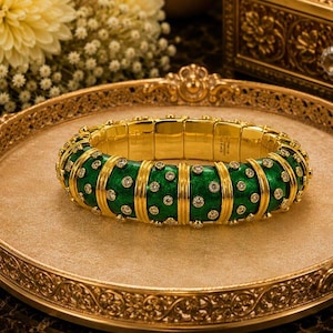 May include: A gold-tone hinged bangle bracelet featuring a vibrant green enamel finish with embedded clear round stones. The bracelet is constructed with alternating vertical gold bands and green enamel segments, creating a structured, repeating pattern.