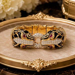 May include: A gold hinged bracelet featuring two panther heads facing each other. The panther heads are decorated with black enamel, clear pavé-set stones, and small red stones for the eyes. A circular cluster of clear stones connects the two heads.