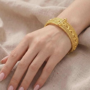 Puede incluir: Una pulsera rígida articulada de tono dorado con un intrincado patrón en relieve de motivos curvos repetitivos y bordes texturizados. La banda de metal ancha y rígida tiene un mecanismo de cierre seguro visible en la parte superior.