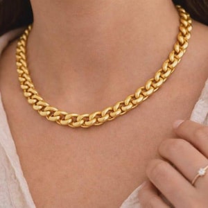 Puede incluir: Un collar de cadena gruesa en tono dorado con eslabones ovalados entrelazados alrededor del cuello, combinado con un anillo fino en tono dorado con una pequeña piedra clara en el dedo. El collar tiene un acabado pulido y un aspecto sólido.