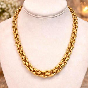 Puede incluir: Un collar de cadena gruesa en tono dorado que presenta una serie de eslabones ovalados y redondeados entrelazados. El collar tiene una superficie pulida y reflectante con un diseño uniforme que se dispone en forma circular sobre el busto.