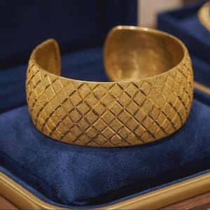 Puede incluir: Una pulsera brazalete ancha de oro con un patrón de diamantes texturizado en toda la superficie exterior. La pulsera tiene un diseño abierto en la parte posterior y un acabado mate, mostrando una cuadrícula geométrica de rombos.