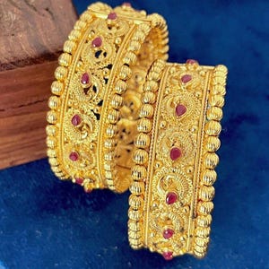 May include: Two gold bangles featuring intricate filigree patterns and small, teardrop-shaped red stones. The edges of the bangles are lined with a textured, beaded design, creating a detailed metallic surface with a polished yellow gold finish.