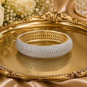 May include: A gold bangle bracelet featuring a wide, rounded band completely encrusted with numerous small, clear, sparkling stones. The interior of the bracelet displays a detailed, repeating geometric honeycomb pattern in polished gold metal.