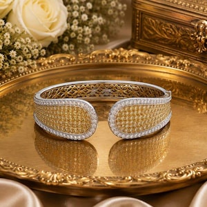 May include: A hinged cuff bracelet featuring a two-tone design with a central panel of pavé-set yellow stones bordered by clear white stones. The interior of the band displays an intricate openwork lattice pattern in a metallic gold finish.