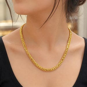 Pode incluir: Um colar de corrente de ouro polido espesso com um padrão denso de elos entrelaçados. O colar tem um acabamento metálico brilhante e reflexivo, assentando rente ao pescoço, exibindo um design uniforme, arredondado e texturizado.