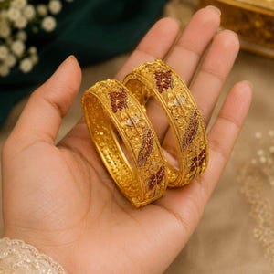 Puede incluir: Dos brazaletes dorados con intrincados trabajos de filigrana, superficies texturizadas e incrustaciones de patrones florales en rojo oscuro y blanco. Los brazaletes tienen un perfil ancho y redondeado con grabados detallados y acabado pulido.