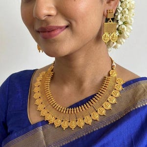 Puede incluir: Un conjunto de collar y pendientes colgantes en tono dorado con una serie de colgantes circulares tipo moneda con figuras en relieve. El collar tiene un diseño de banda ancha texturizada con monedas colgantes y pendientes cuadrados.