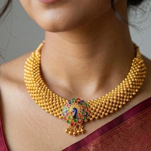 Puede incluir: Un collar tipo gargantilla dorado con una cadena de cuentas texturizadas y un colgante central en forma de pavo real. El colgante está decorado con esmalte vibrante en tonos azul, verde y rojo, con pequeñas cuentas doradas colgando.
