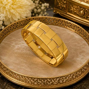 May include: A wide, polished gold bracelet featuring a repeating woven or basket-weave pattern of interlocking rectangular links. The bracelet has a high-shine, reflective metallic finish and a structured, rigid circular shape.