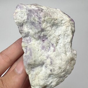 May include: A natural fluorite specimen with a rough texture. The stone displays a mix of white and pale purple colours. The mineral is held in a hand against a plain background.