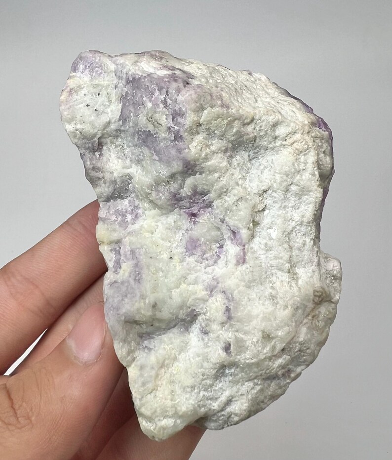 May include: A natural fluorite specimen with a rough texture. The stone displays a mix of white and pale purple colours. The mineral is held in a hand against a plain background.