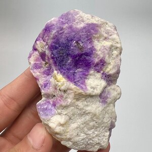 May include: A natural fluorite specimen with a raw, unpolished appearance. The stone displays a vivid contrast of deep purple and white colours. The crystalline structure is visible, with a rough texture. The mineral is held in a hand against a plain background.