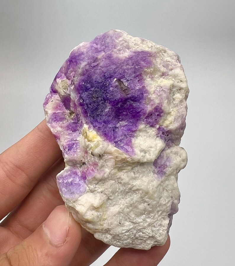 May include: A natural fluorite specimen with a raw, unpolished appearance. The stone displays a vivid contrast of deep purple and white colours. The crystalline structure is visible, with a rough texture. The mineral is held in a hand against a plain background.
