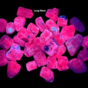 Puede incluir: Un conjunto de rocas rosas y moradas brillantes sobre un fondo negro. Las rocas tienen una textura rugosa y formas variadas. Algunas rocas tienen manchas azules y moradas oscuras. El texto "Long Wave" está en la esquina superior izquierda.