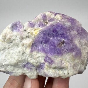 May include: A natural mineral specimen with a mix of colours. The stone shows a prominent area of deep purple, contrasted by patches of white and hints of yellow. The rough texture and irregular shape highlight its natural origin.