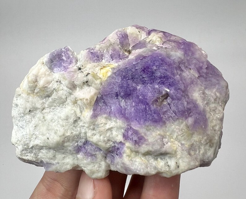 May include: A natural mineral specimen with a mix of colours. The stone shows a prominent area of deep purple, contrasted by patches of white and hints of yellow. The rough texture and irregular shape highlight its natural origin.