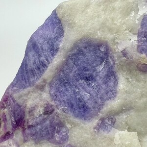 May include: Close-up of a natural stone with a mix of purple and white hues. The stone features large, rounded purple sections set against a backdrop of lighter, off-white areas. The texture appears rough and crystalline.