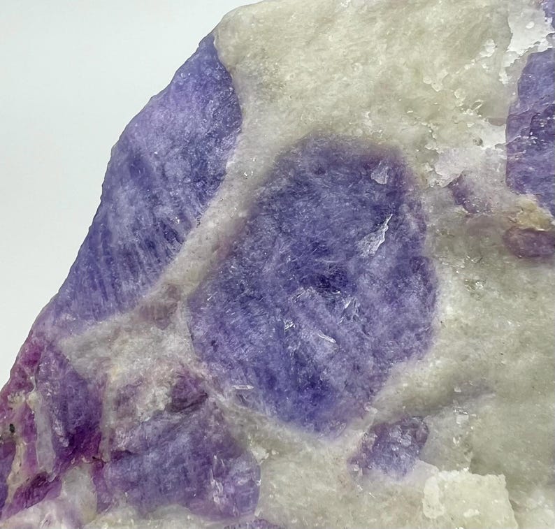 May include: Close-up of a natural stone with a mix of purple and white hues. The stone features large, rounded purple sections set against a backdrop of lighter, off-white areas. The texture appears rough and crystalline.