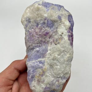 May include: A natural mineral specimen with a mix of colours. The stone features light purple, white, and pink hues. The rough texture and natural form of the mineral are visible. The mineral is held in a hand.