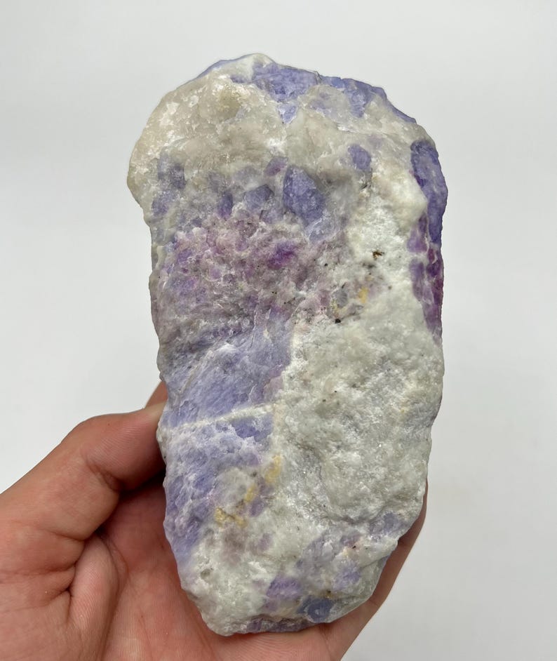 May include: A natural mineral specimen with a mix of colours. The stone features light purple, white, and pink hues. The rough texture and natural form of the mineral are visible. The mineral is held in a hand.