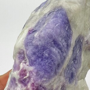 May include: Close-up of a natural stone with a mix of colours. The stone features large patches of deep purple and lavender hues, contrasted by areas of white and pale pink. The texture appears crystalline and rough.