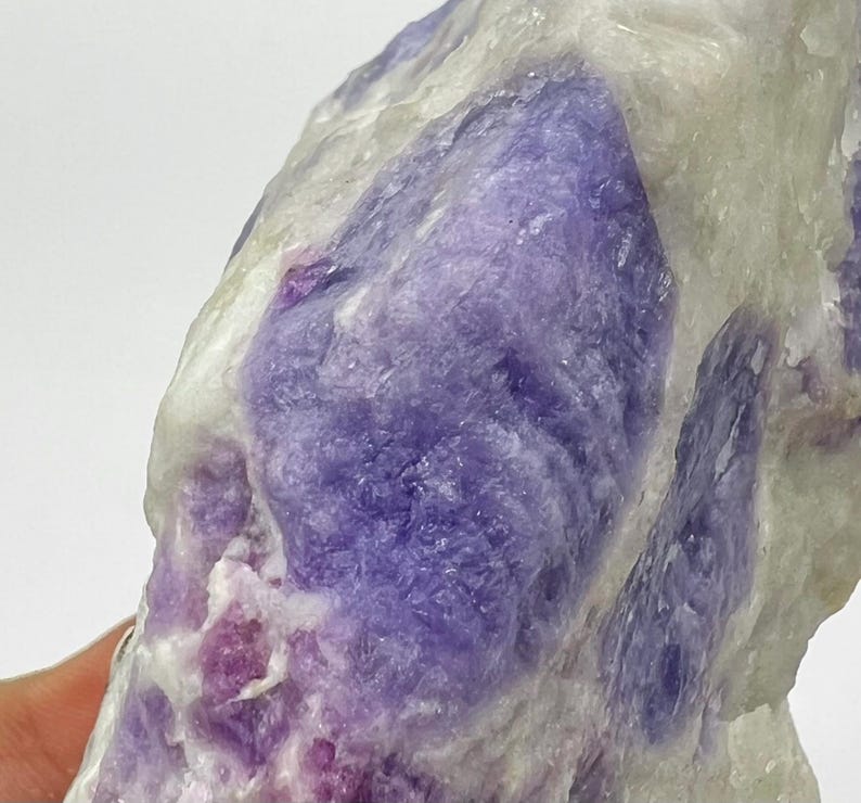 May include: Close-up of a natural stone with a mix of colours. The stone features large patches of deep purple and lavender hues, contrasted by areas of white and pale pink. The texture appears crystalline and rough.