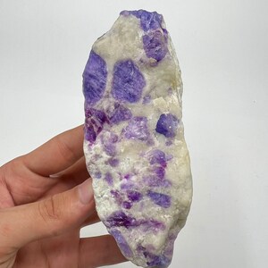 May include: A natural gemstone with a mix of white and purple colours. The stone has a rough, irregular shape and is held in a hand. The purple sections are clustered throughout the white base, creating a unique pattern. This is a mineral specimen.
