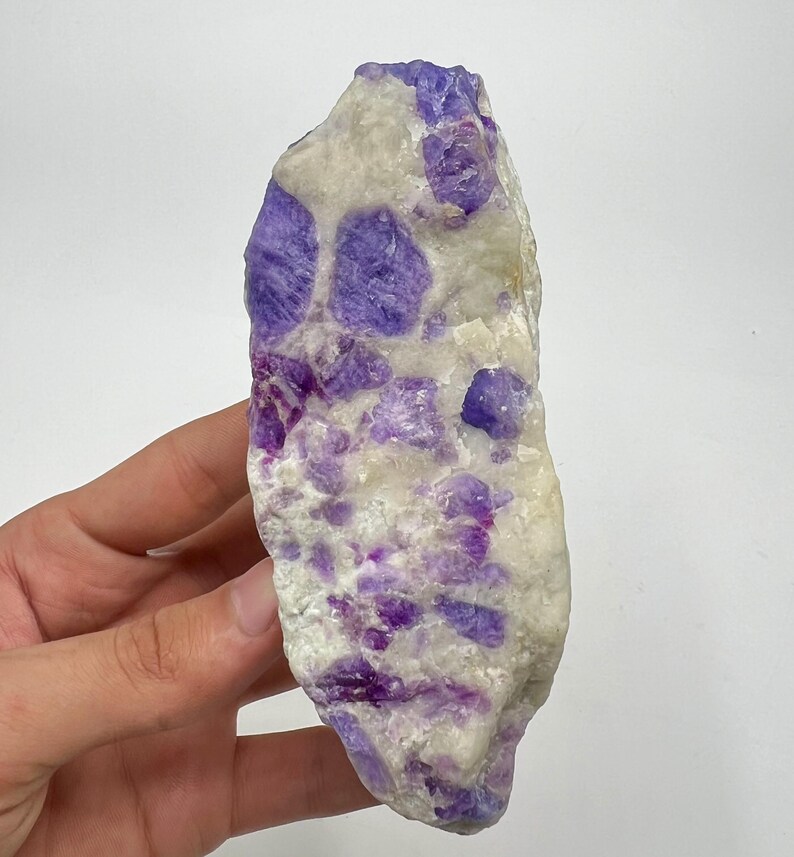 May include: A natural gemstone with a mix of white and purple colours. The stone has a rough, irregular shape and is held in a hand. The purple sections are clustered throughout the white base, creating a unique pattern. This is a mineral specimen.