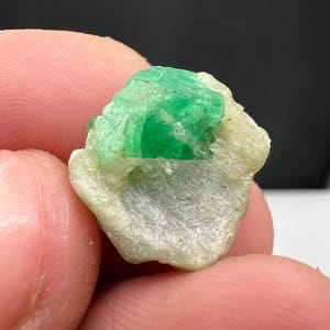 May include: A raw emerald gemstone with a vibrant green hue, set within a light-colored matrix. The rough, uncut stone displays a natural, unpolished texture, showcasing its crystalline structure. The emerald is held between fingers.