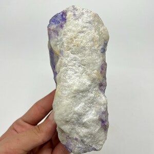 May include: A natural fluorite crystal. The stone has a rough, textured surface with a mix of white and purple colours. The crystal is held in a hand against a white background.