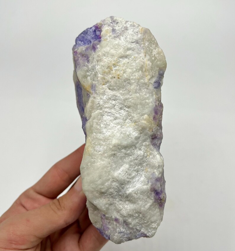 May include: A natural fluorite crystal. The stone has a rough, textured surface with a mix of white and purple colours. The crystal is held in a hand against a white background.