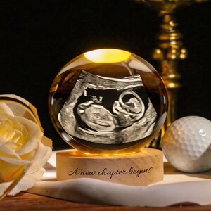 Könnte beinhalten: Eine leuchtende Glaskugel mit einem Ultraschallbild eines Babys im Inneren, auf einem Holzsockel. Auf dem Sockel steht "A new chapter begins". Eine gelbe Rose, ein Golfball und andere dekorative Gegenstände umgeben die Kugel.