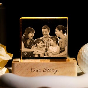 May include: A sepia-toned family photo is displayed within a clear, illuminated glass block. The block sits on a wooden base engraved with "Our Story." A golf ball and a decorative flower are in the foreground.
