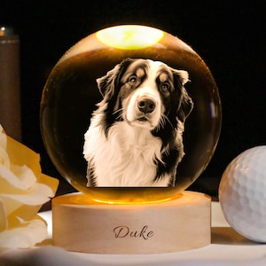 May include: A glass sphere featuring a detailed dog image, set on a wooden base. The base is lit with a warm light, and the name "Duke" is inscribed. A golf ball is positioned beside the sphere, with a dark background.