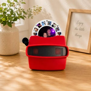 Pode incluir: Um visualizador estereoscópico de plástico vermelho brilhante com um disco branco circular inserido na ranhura superior. O disco exibe várias pequenas imagens fotográficas, incluindo uma foto central de duas pessoas. O dispositivo tem uma área de lente preta.